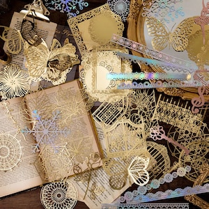 May include: Assortment of gold and iridescent decorative metal stickers featuring butterflies, snowflakes, and intricate patterns. The stickers are arranged on a book and a wooden surface.
