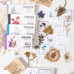 May include: A collection of decorative paper ephemera, including small notebooks with floral illustrations and Japanese text, dried flowers, and vintage-style cards. The color palette includes white, beige, and pastel hues, creating a delicate and artistic aesthetic.
