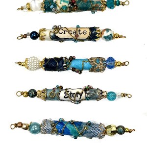 May include: Five blue and white beaded jewelry pieces with gold wire accents. The beads are wrapped with fabric and have words on them, including "Create" and "Story".