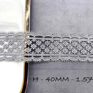 May include: A silver metallic lace trim featuring a repeating floral pattern with scalloped edges. The lace has a height of 40 mm or 1.57 inches as indicated by the text printed on the image, showcasing a detailed woven texture.