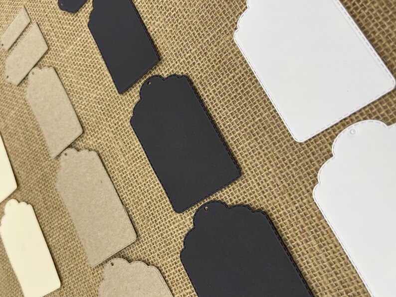 May include: Assortment of blank gift tags in various colors and materials, including white, black, and brown. The tags have a scalloped edge and a hole for string or ribbon.