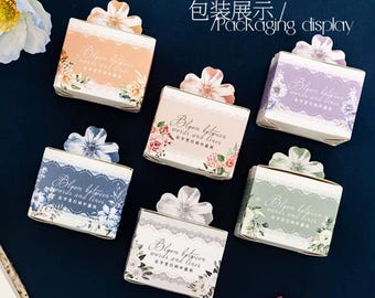 May include: Six rectangular paper boxes in orange, purple, blue, pink, green, and white, each featuring a floral cutout tab on top. The boxes are decorated with lace patterns and floral illustrations, with the text Bloom between words and lines.