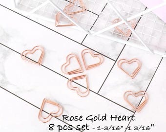 May include: A set of eight rose gold heart-shaped paper clips, each measuring 1-3/16 inches by 1-3/16 inches. The metal clips are arranged in a scattered pattern, with some resting near a white wire grid organizer on a white marble surface.
