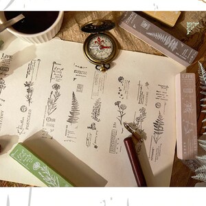 May include: A set of 10 clear rubber stamps featuring floral and fern designs. The stamps are arranged on a white paper background with a compass, a pen, and other stamps. The stamps include words such as "Life", "Imagine", "Healthy", and "Postcard".