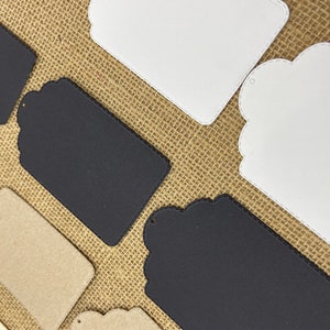 May include: Assortment of blank gift tags in various shapes and sizes. The tags are made of different materials, including white cardstock, black cardstock, and brown kraft paper. Some tags have scalloped edges and a hole for string.