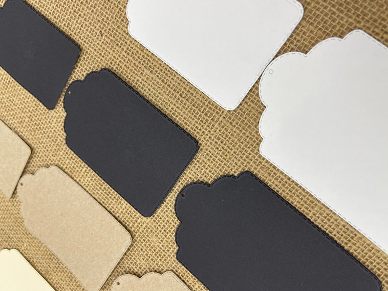 May include: Assortment of blank gift tags in various shapes and sizes. The tags are made of different materials, including white cardstock, black cardstock, and brown kraft paper. Some tags have scalloped edges and a hole for string.
