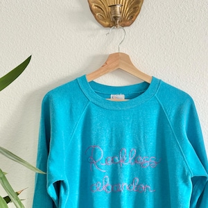 May include: A turquoise blue sweatshirt with the embroidered words "Reckless abandon" in pink script. The long-sleeved top is hanging on a wooden hanger against a white wall. A gold-coloured wall sconce is above the hanger.