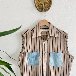 May include: Sleeveless shirt with brown vertical stripes on a beige background. It has two light blue denim pockets and the text "ELYSIAN FIELDS" on the label. The shirt is suspended from a wooden hanger.