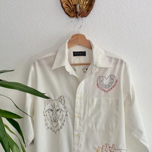 May include: White button-down shirt with embroidered designs. Features a wolf head, a heart with text, and a hand holding a flame. The shirt has a pocket and is hanging on a wooden hanger.