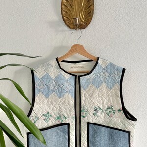 May include: A sleeveless quilted vest featuring a patchwork design of light blue, white, and floral fabric squares. The vest has two large blue denim-like patch pockets, a round neckline, and black trim. The interior label reads ELYSIAN FIELDS.