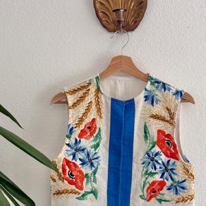 May include: A cream-colored vest with embroidered floral designs. The vest features red poppies, blue flowers, and wheat stalks, with a blue border and vertical stripes. It hangs on a wooden hanger.