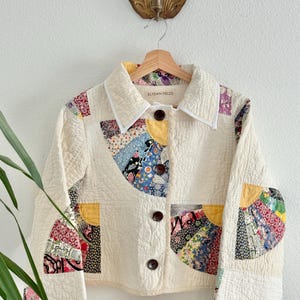 May include: A cropped jacket made from cream-colored quilted fabric featuring colorful fan-shaped patchwork panels in shades of yellow, blue, red, and black. The jacket has a pointed collar with white trim, four dark brown buttons, and a label reading ELYSIAN FIELDS.