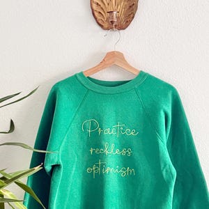 May include: A green sweatshirt with the words "Practice reckless optimism" embroidered in a cursive font. The sweatshirt is hanging on a wooden hanger against a white wall. A gold-coloured wall decoration is above the sweatshirt.