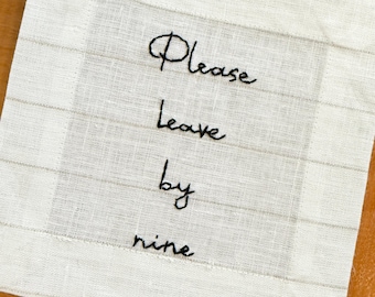 Puede incluir: Una servilleta cuadrada de lino blanquecino con el texto bordado "Please Leave by nine" en hilo negro. La servilleta tiene un sutil patrón de cuadrícula y un diseño simple y elegante.
