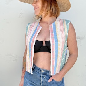 Könnte beinhalten: Eine ärmellose Weste mit pastellfarbenem Streifenmuster in Rosa, Blau, Gelb und Grün. Die Weste wird mit Jeansshorts und einem Strohhut getragen. Das Outfit hat eine Vintage- und Retro-Ästhetik.