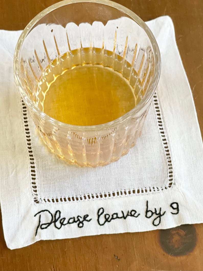 K&ouml;nnte beinhalten: Eine wei&szlig;e Leinen-Serviette mit schwarzer Stickerei, auf der "Please leave by 9" steht. Ein klares Glas mit einer goldenen Fl&uuml;ssigkeit steht auf der Serviette.
