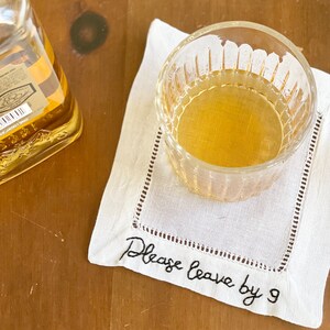 K&ouml;nnte beinhalten: Ein wei&szlig;er Leinen-Untersetzer mit schwarzer Stickerei, auf dem "Please leave by 9" steht. Ein Glas mit bernsteinfarbener Fl&uuml;ssigkeit steht auf dem Untersetzer.