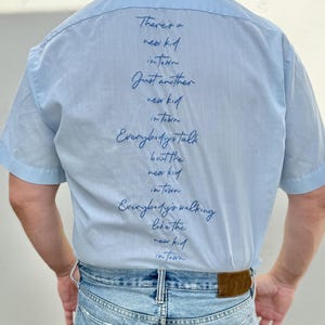 May include: Pale blue short-sleeved shirt with dark blue script text including "There's a new kid in town". The person is wearing light blue denim jeans and a brown leather belt.