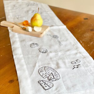 May include: White linen table runner with black embroidered images of a pear, cheese, strawberries, olives, and honey.