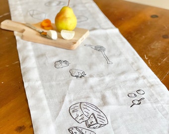 Può includere: Un runner da tavolo in lino bianco con immagini ricamate in nero di una pera, formaggio, fragole, olive e miele.