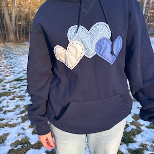 May include: Navy blue hoodie with a front pocket and a drawstring hood. The hoodie features three heart-shaped patches in white, light blue floral, and blue. The patches are stitched onto the front of the hoodie.