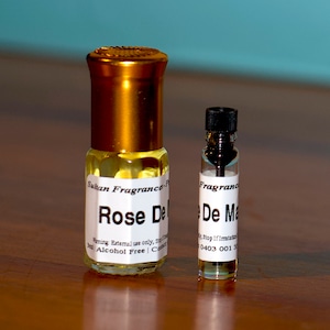 Op de afbeelding: Twee glazen flessen parfum, een groter dan de andere. De grotere fles is goudkleurig met een wit etiket waarop "Rose De... Sultan Fragrance... Alcoholvrij" staat. De kleinere fles is helder met een zwarte dop en een wit etiket waarop "De Ma... Fragrance" staat.