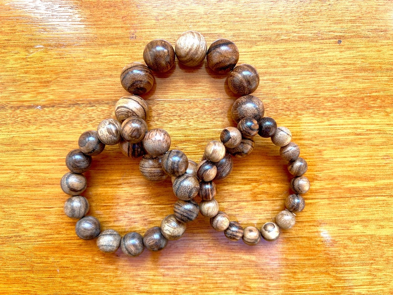 May include: Three wooden bracelets with brown and black beads. The bracelets are arranged in a circle on a wooden surface.