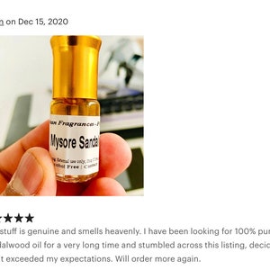 Op de afbeelding: Een klein, doorzichtig glazen flesje met een gouden dop en een etiket met de tekst "Mysore Sandal" en "Alcohol Free". Het flesje wordt vastgehouden door een hand.