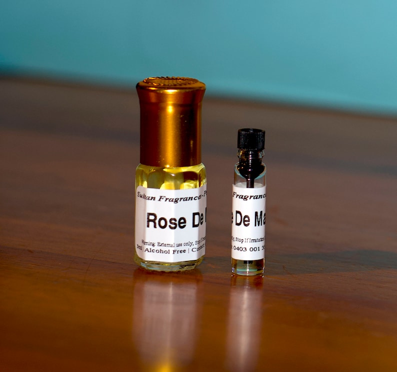 Op de afbeelding: Twee glazen flessen parfum, een groter dan de andere. De grotere fles is goudkleurig met een wit etiket waarop "Rose De... Sultan Fragrance... Alcoholvrij" staat. De kleinere fles is helder met een zwarte dop en een wit etiket waarop "De Ma... Fragrance" staat.