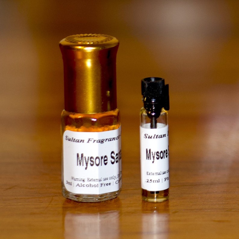 Op de afbeelding: Twee glazen flessen parfum met gouden doppen en witte etiketten. De kleinere fles is gelabeld "Sultan Mysore 25ml Ph" en de grotere fles is gelabeld "Sultan Fragrand Mysore Sandal 3ml Alcohol Free".