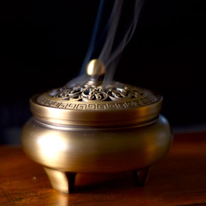 Op de afbeelding: Een gouden metalen wierookbrander met een decoratief deksel en drie poten. Rook stijgt op uit de brander.