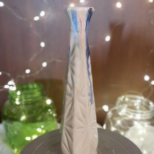 Pode incluir: Um vaso alto de cerâmica branca com um padrão geométrico e uma parte superior alargada. A parte superior do vaso tem pingos de tinta azul, rosa e branca. O vaso está sobre uma superfície cinza, com um fundo desfocado de luzes de fadas e potes.