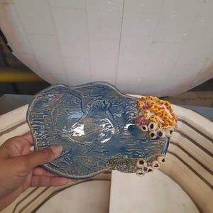 May include: A handmade ceramic plate featuring a whale design. The plate is light blue with raised relief detailing of whales and ocean waves. The edge has a textured, coral-like design with red, yellow, and white accents.