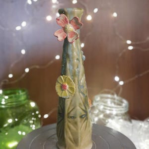 May include: A handmade ceramic vase with a floral design. The vase is primarily green with blue and brown accents. It features a pink and yellow flower embellishment near the top and a yellow flower on the side. The vase is on a metal surface.