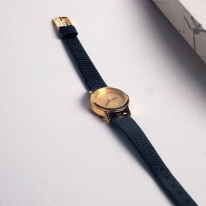 May include: A gold-toned wristwatch with a dark green textured leather strap. The watch face is gold with simple markers. The buckle is gold. The watch is displayed on a white surface.
