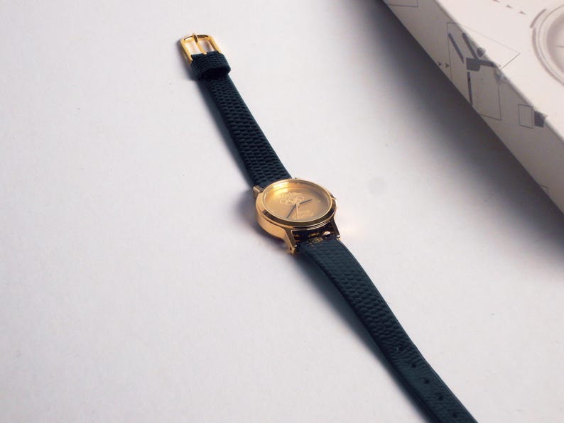 May include: A gold-toned wristwatch with a dark green textured leather strap. The watch face is gold with simple markers. The buckle is gold. The watch is displayed on a white surface.