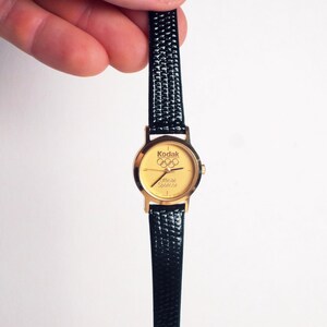 May include: Gold-toned wristwatch with a black textured band. The watch face features the Kodak logo and Olympic rings, with the text "Official Sponsor." The watch is held against a white background.