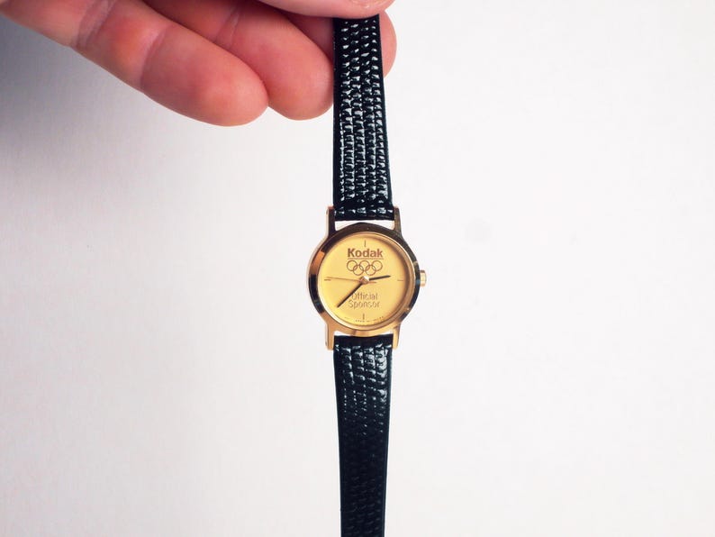 May include: Gold-toned wristwatch with a black textured band. The watch face features the Kodak logo and Olympic rings, with the text "Official Sponsor." The watch is held against a white background.