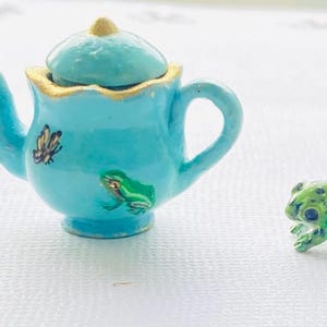 May include: A miniature turquoise teapot with a gold-trimmed rim and lid, adorned with a butterfly and a green frog. Beside it is a small green frog figurine. The teapot is a charming decorative item.