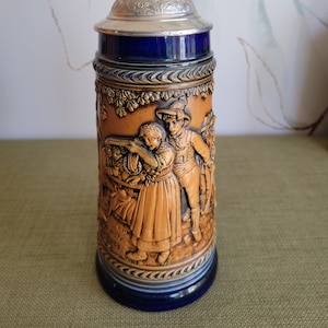 May include: A ceramic beer stein with a blue and brown design. The stein features a relief sculpture of a couple in traditional German clothing. The lid is silver with a decorative design.