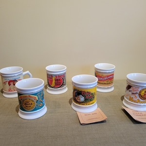 May include: Six vintage-style ceramic mugs with various advertising designs. The mugs feature images of a woman, a giraffe, a rooster, a bird, a squirrel, and a man. The mugs are all white with gold trim.