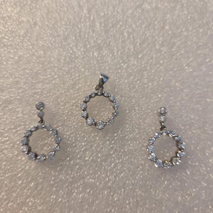 May include: A jewelry set featuring a pendant and a pair of earrings. The pendant and earrings have a circular design, each adorned with multiple small, sparkling stones. The set is made of a silver-colored metal.