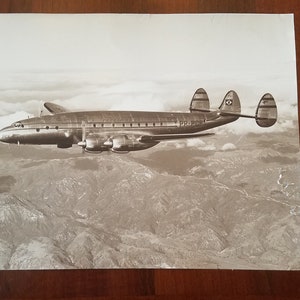 May include: A black and white photograph of a Lockheed Constellation airplane flying over a mountainous landscape. The airplane has three tail fins and the text "PP-PBR" is visible on the side of the fuselage.
