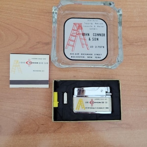 May include: A vintage lighter with a silver finish and a yellow plastic insert. The lighter features a white label with a red and black logo for John Connor & Son, a ladder company in Rochester, New York. The label also includes the company's phone number and address.