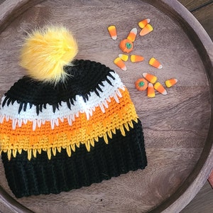 May include: A black, orange, white, and yellow crocheted beanie with a fluffy yellow pom pom on top. The beanie is sitting on a wooden surface with candy corn scattered around it.