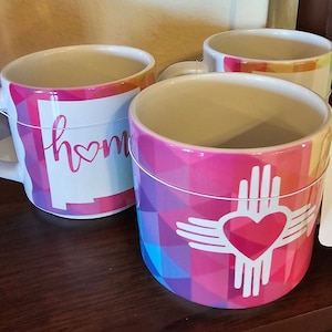 May include: Three ceramic mugs with colorful geometric designs. One mug says "home" with a heart. Another mug has a white heart inside a stylized Zia symbol.