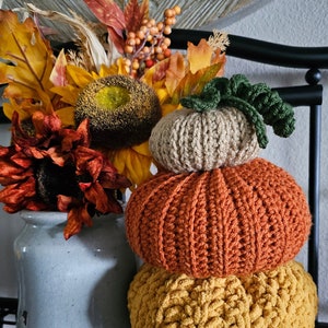 May include: A stack of three crocheted pumpkins in fall colors. The bottom pumpkin is yellow, the middle pumpkin is orange, and the top pumpkin is beige. The pumpkins are decorated with a green crocheted stem.