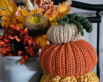 May include: A stack of three crocheted pumpkins in fall colors. The bottom pumpkin is yellow, the middle pumpkin is orange, and the top pumpkin is beige. The pumpkins are decorated with a green crocheted stem.