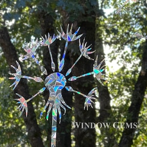 May include: A holographic dandelion window cling with a white silhouette and iridescent colors. The cling is placed on a window with a view of trees and foliage.