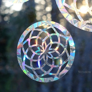 May include: A set of iridescent, geometric window clings in the shape of a flower. The clings are made of a thin, reflective material and have a rainbow-like appearance. The text "WINDOW GEMS™" is visible in the bottom right corner of the image.